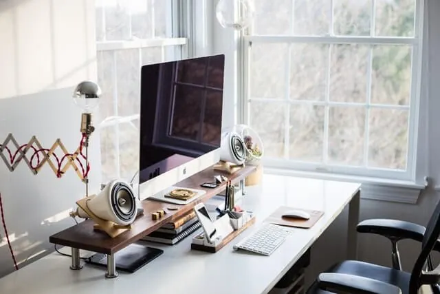 A workspace with a large screen and task lamp beside bright windows.