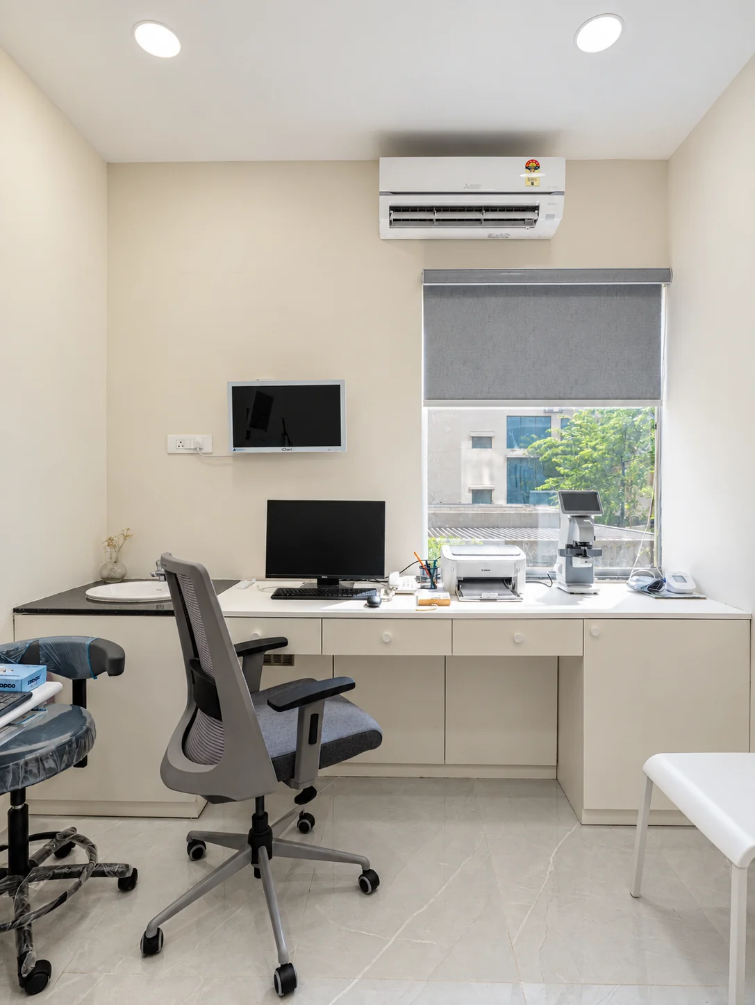 Clinic consultation room with a fitted white desk, storage cabinets and a window with a roller blind.